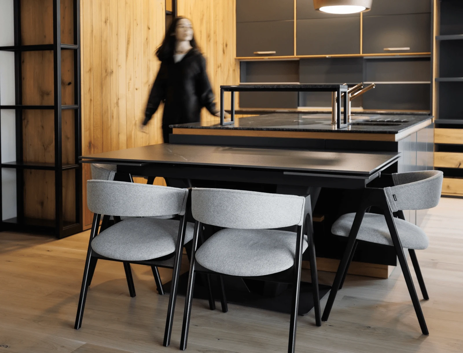 A woman walking past the kitchen island with four grey-fabric Lain chairs on black-stained ash frames around a dark dining table