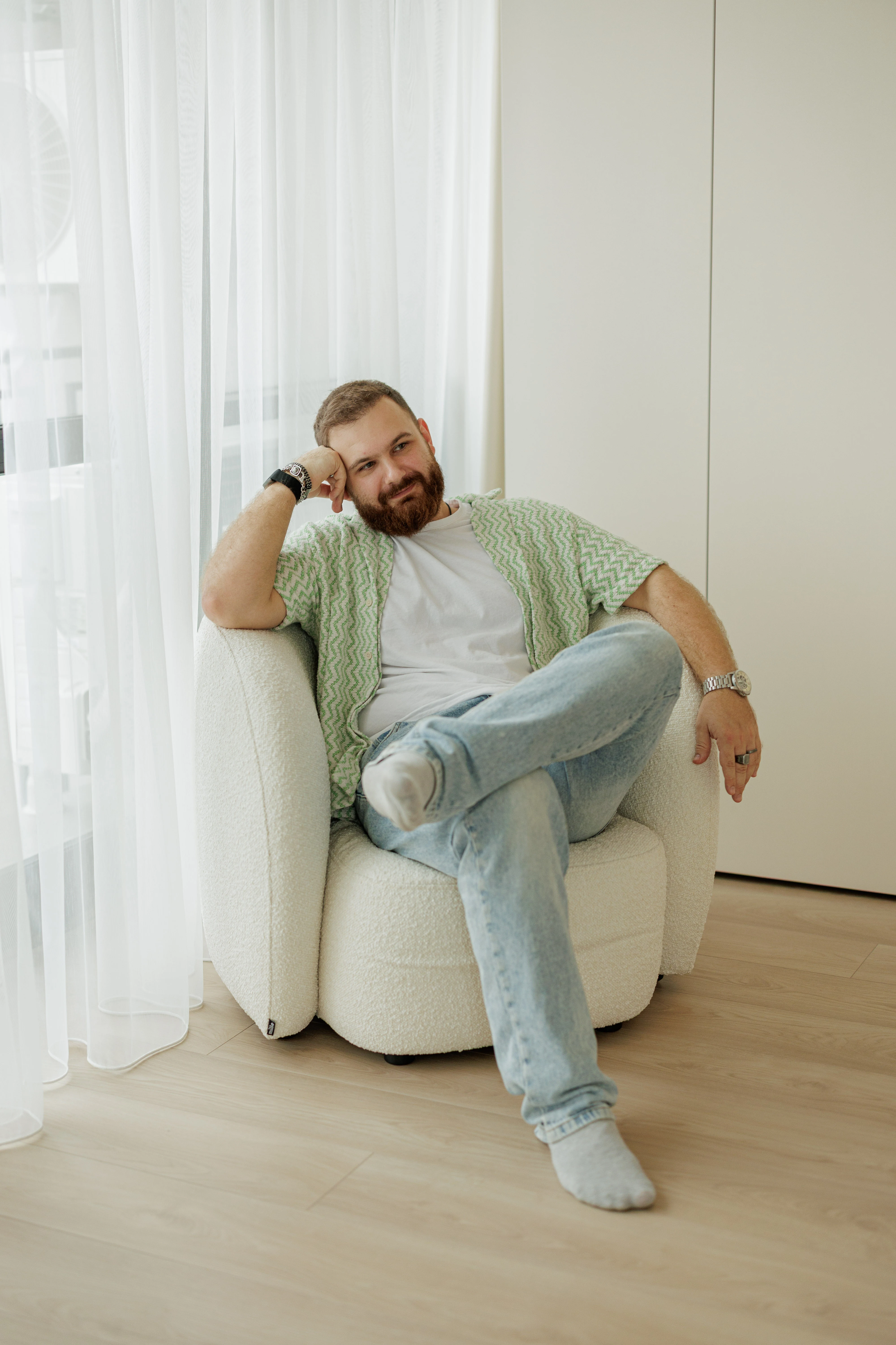 A man in a green-and-white striped shirt and jeans seated in a cream boucle Nuba armchair, hand to his cheek, against a wall and sheer curtain