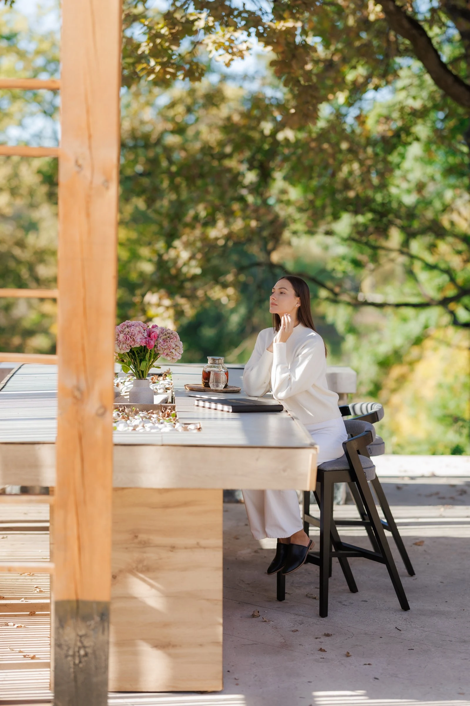 Model in cream seated at an outdoor stone counter with two Baffi bar stools, autumn trees and hydrangeas on the counter