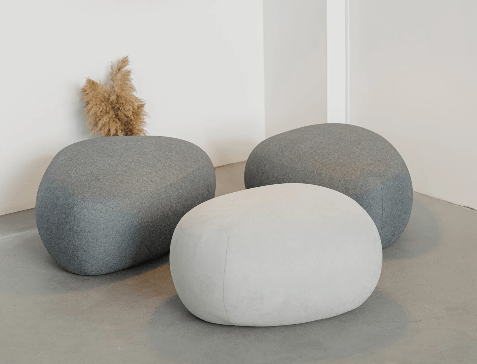 Three Chios poufs in cream, grey and ivory fabrics on a polished cement studio floor with pampas grass behind