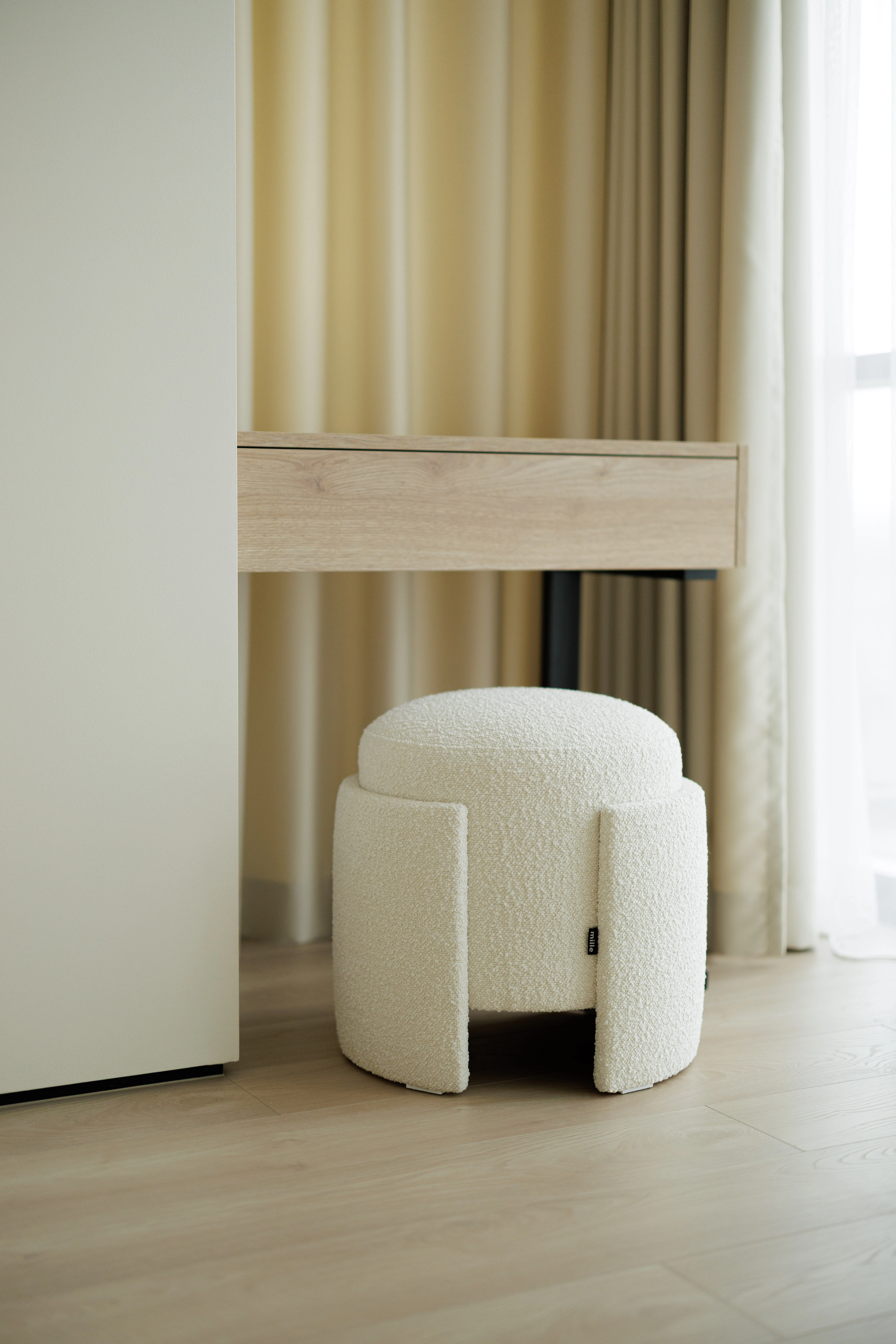 Cream bouclé Eden pouf tucked under a wood vanity desk with gold curtains