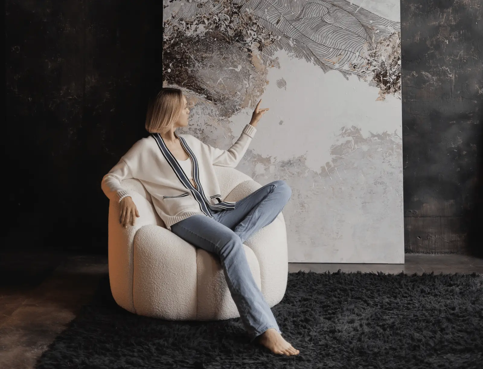 Model seated on the cream Flora chair in a moody dark-walled room with abstract framed artwork