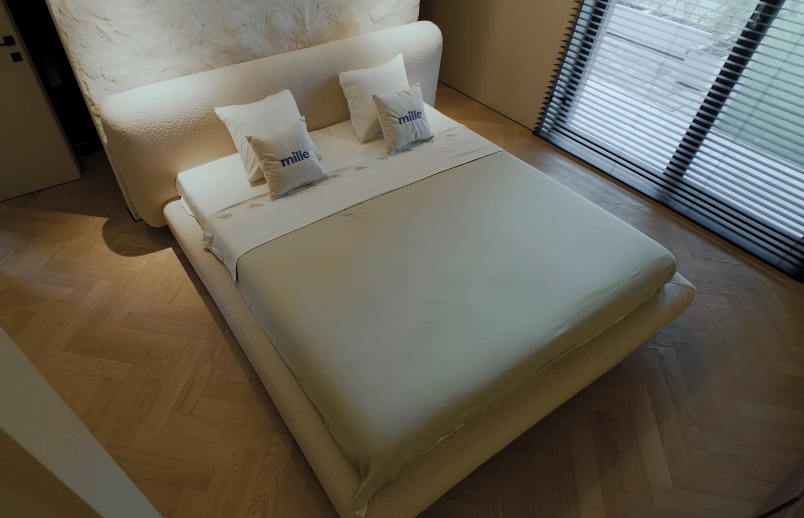 Cream Fresco bed photographed from above in a textured plaster-wall bedroom with dark venetian blinds and herringbone parquet