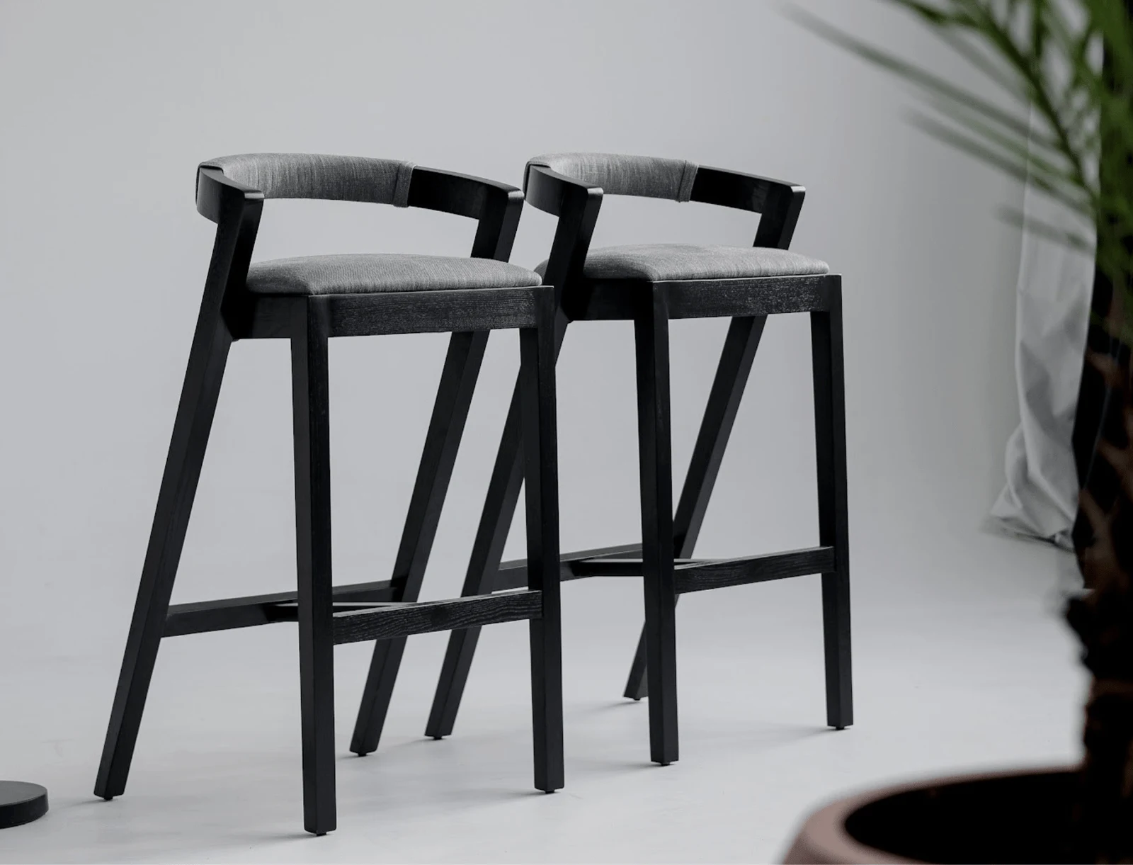 Two Baffi bar stools on a clean white studio floor, black-stained ash sawhorse frames and grey upholstered seats