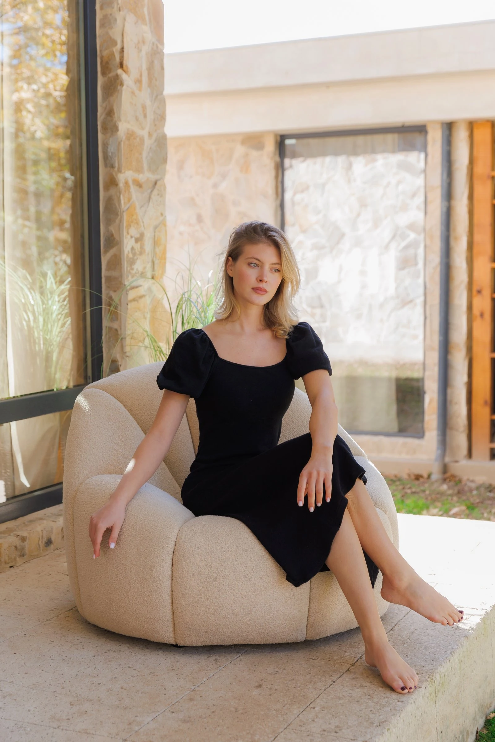 Model in a black dress seated on a cream Flora chair on a stone-walled villa patio with golden autumn light