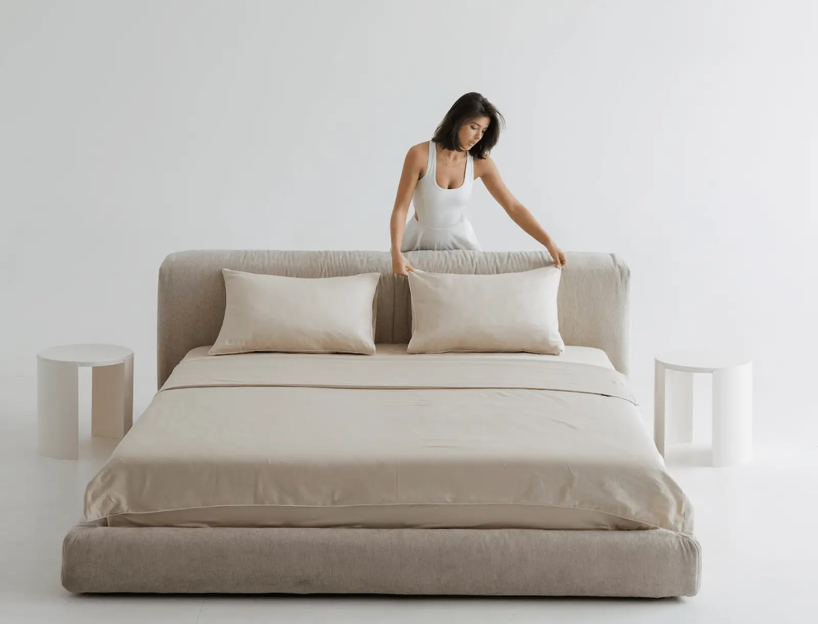 A woman in a white slip dress arranging pillows on a sand Marmaris in a bright minimal studio