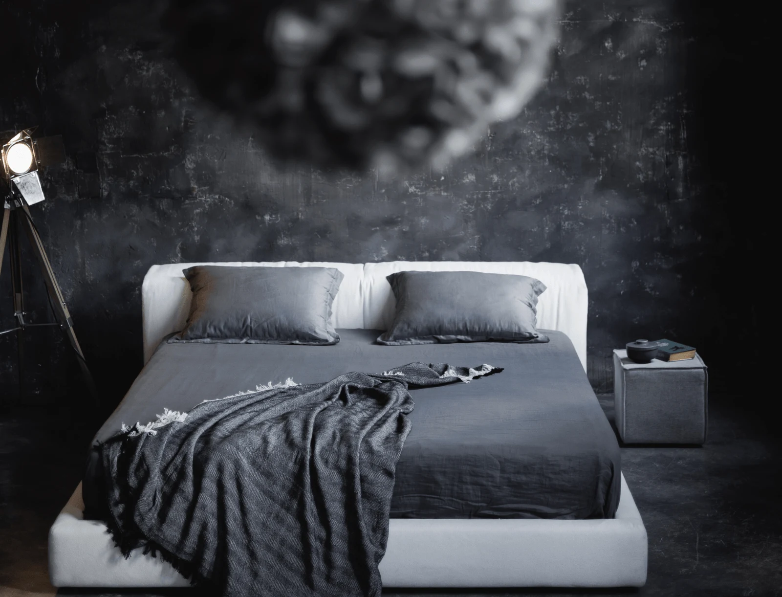 Cream Marmaris in a dark loft with a grey throw draped diagonally across the bed and a tripod light at left