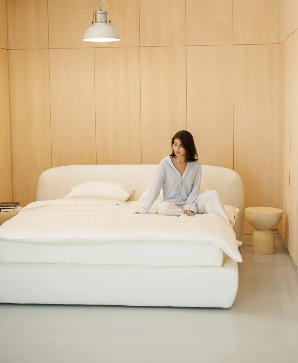 Cream boucle Belagio with a model in pale blue pajamas sitting on the bed, wood-paneled bedroom