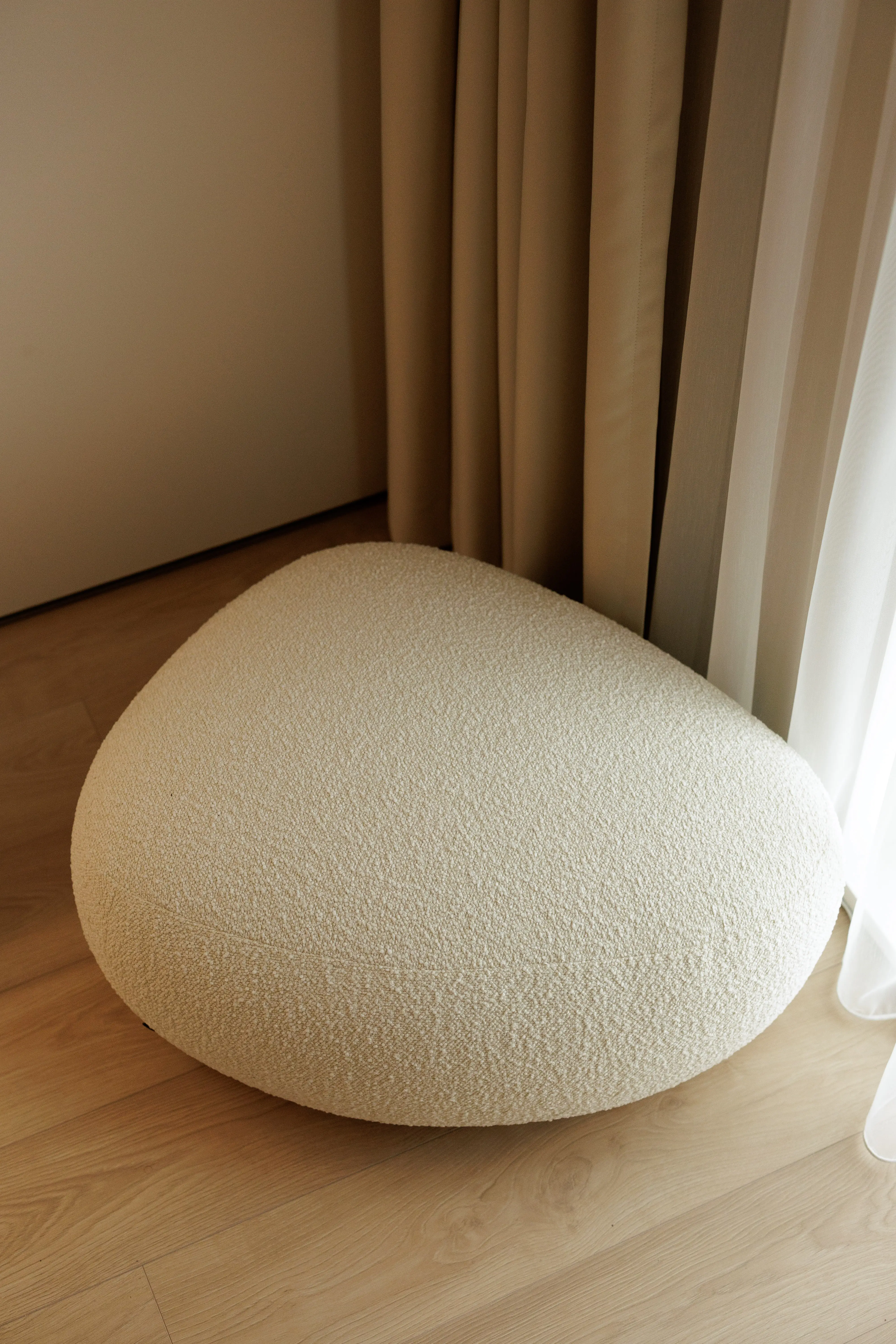 Cream bouclé Chios pouf beside a curtained window on a wood floor
