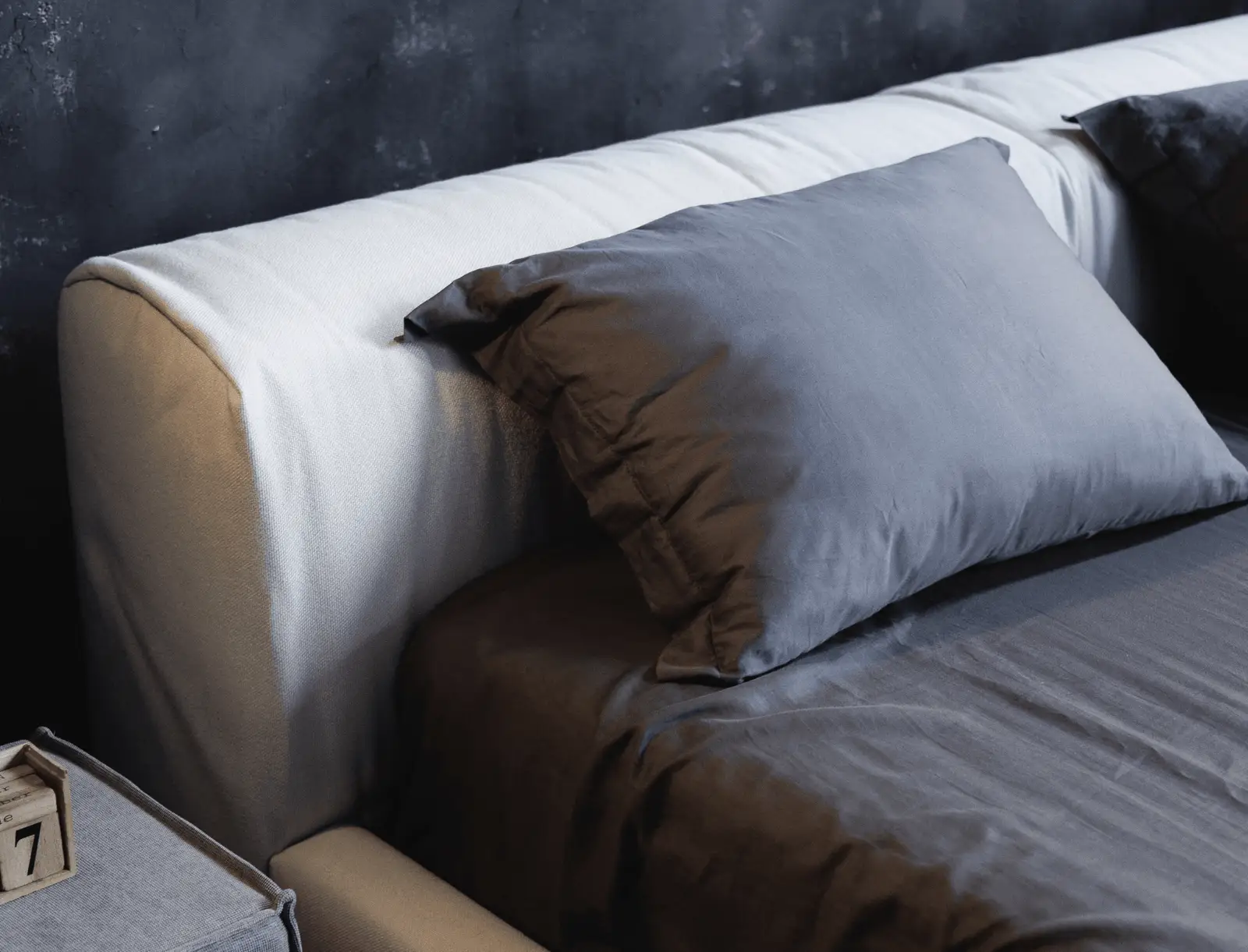Macro of the Marmaris cream headboard with a brown silk pillow and a grey pillow against a dark concrete wall