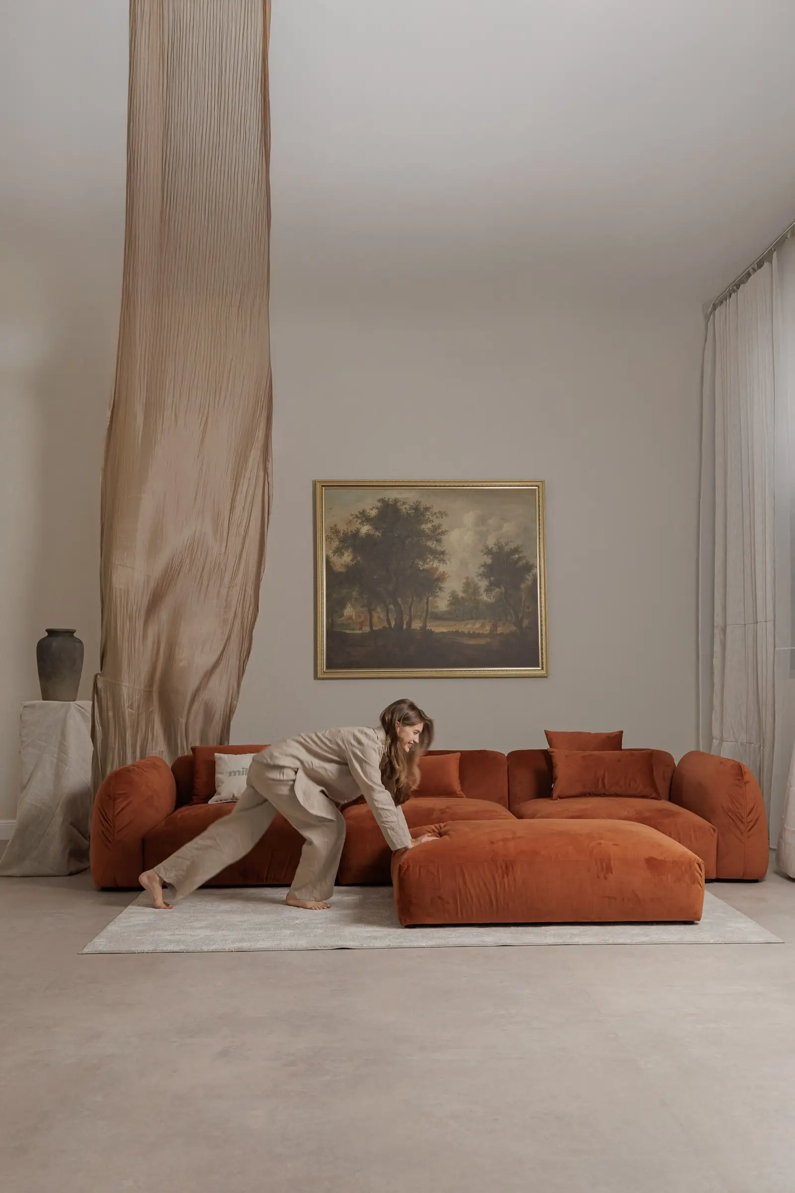 Rust velvet Tesa with model in linen reaching for the ottoman, gilt-framed landscape painting on the wall, sheer curtain behind