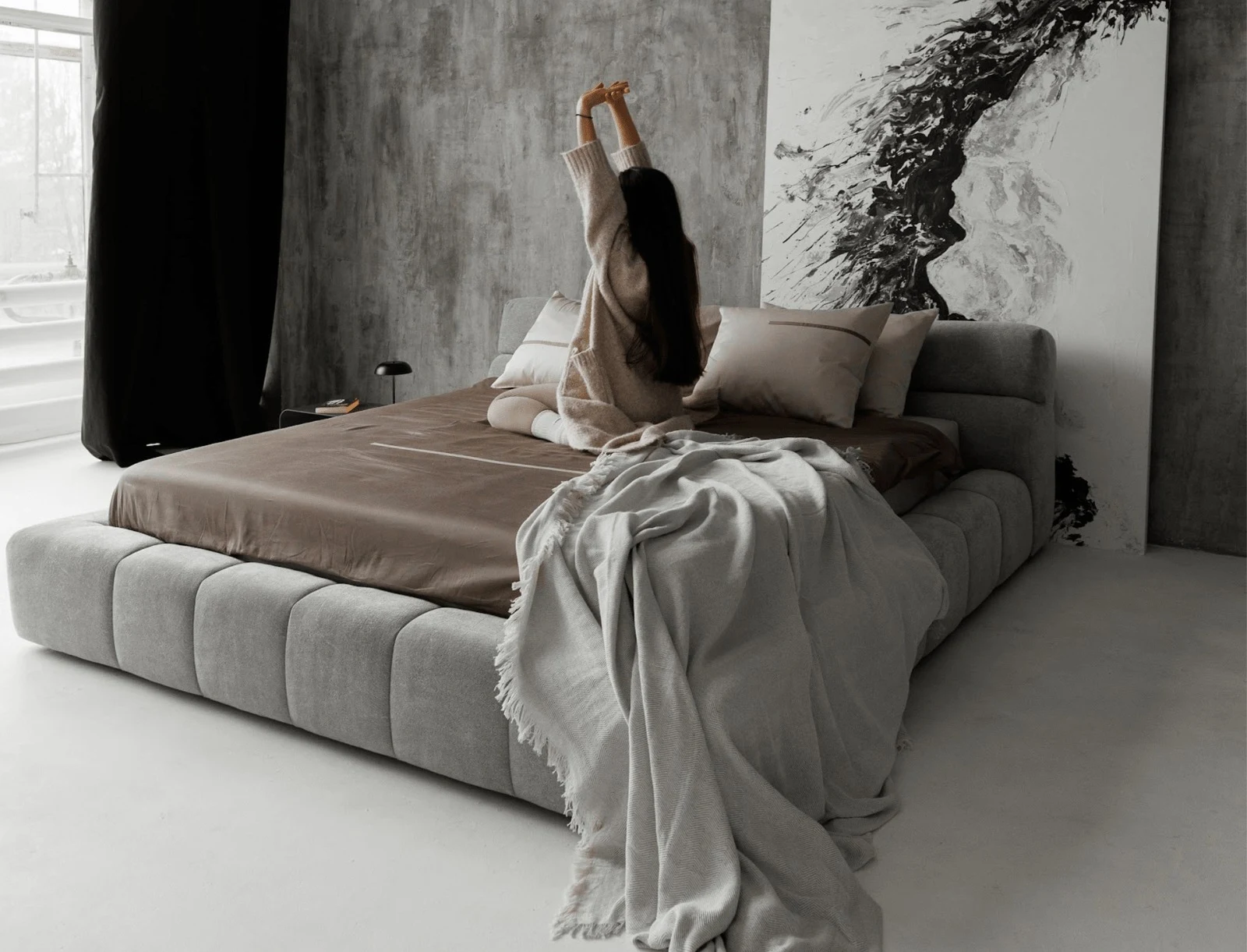 Mid-grey Belgian velvet Nao bed in a concrete-walled bedroom with a model stretching upward