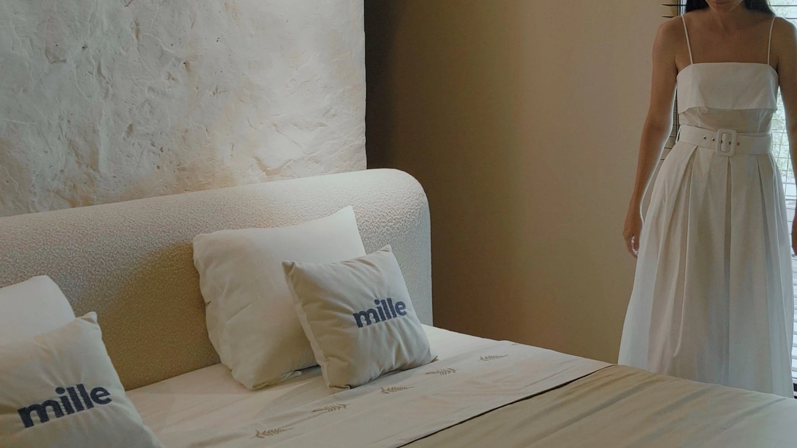 Close on Mille-branded pillows arranged on the cream Fresco bed with a woman in a white dress walking past