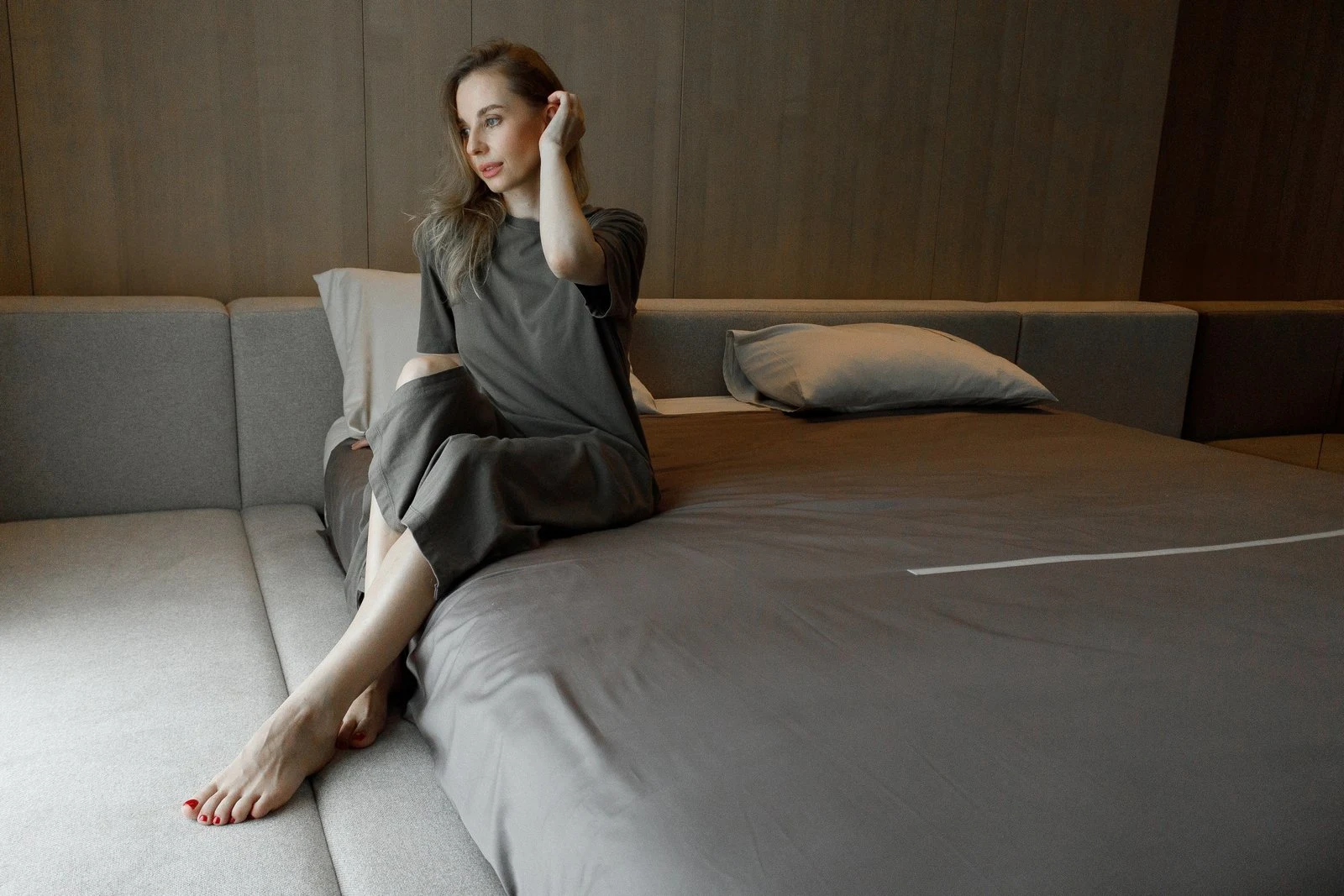 A woman in a dark linen top and skirt sitting on the side bench of a sand Connect bed in a wood-paneled bedroom