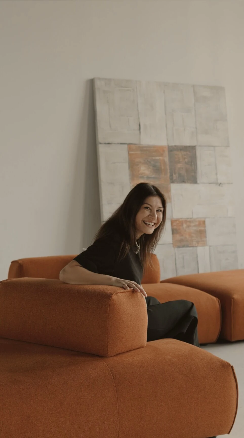 Model smiling on the rust Nissa after rearranging the backrest, with an abstract art panel behind